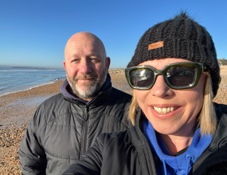 Head and shoulders shot of smiling woman in sunglasses and beanie. Man stands behind her as they stand on a beach